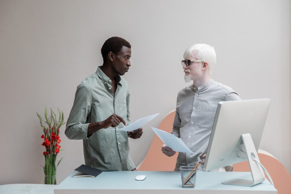 Focused African American man and albino male colleague standing near table with computer and documents in hands while discussing work and looking at each other