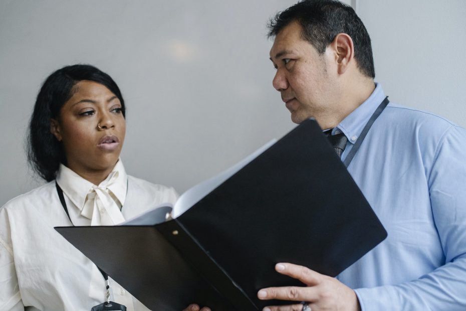 Serious middle aged male manager in shirt showing folder with documents to attentive female African American colleague during work in modern office