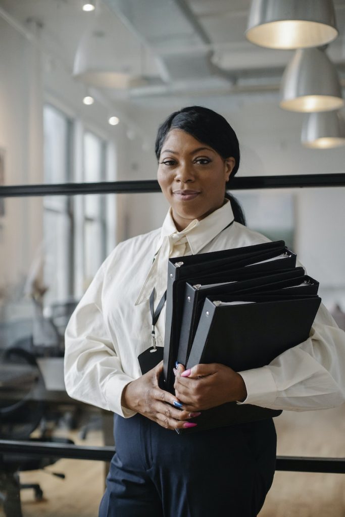 Smiling African American female employee wearing classy outfit standing with paper documents in modern workplace