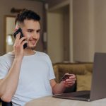 A Man Holding a Credit Card Using a Cellphone in Front of a Laptop