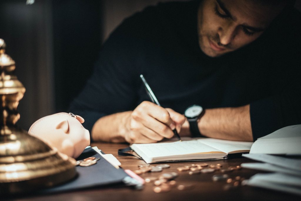 Crop elegant man taking notes in journal while working at desk with coins and piggybank in lamplight